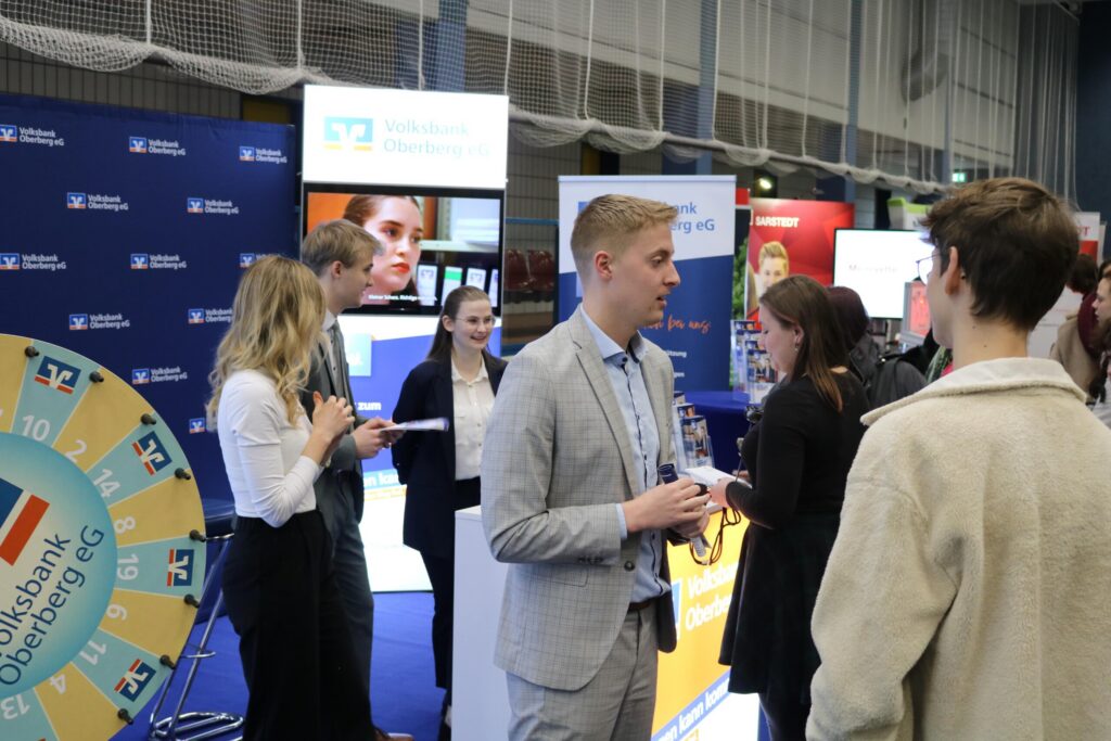 Menschen stehen an einem Messestand und sprechen miteinander. Verschiedene Informationsstände sind im Hintergrund.