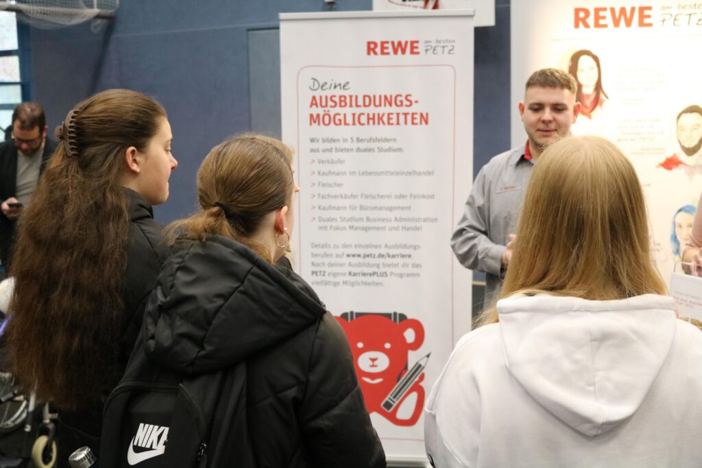Schülerinnen hören einem REWE-Mitarbeiter am Informationsstand zu.