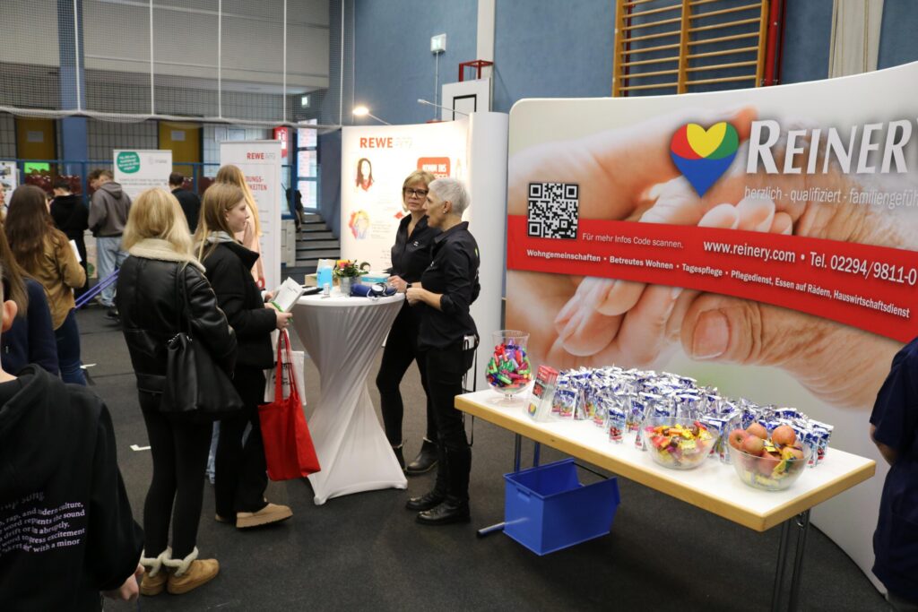 Messestand mit Personen, Informationsmaterial und Snacks in einer Halle.