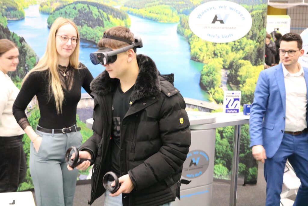 Junge mit VR-Brille und Controllern, umgeben von Messepersonal und Werbung.