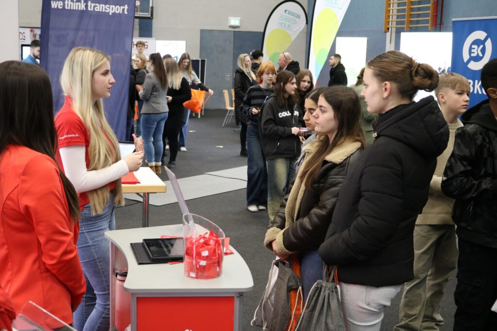 Messe mit mehreren Ständen und Besuchern, eine Person berät Interessierte.