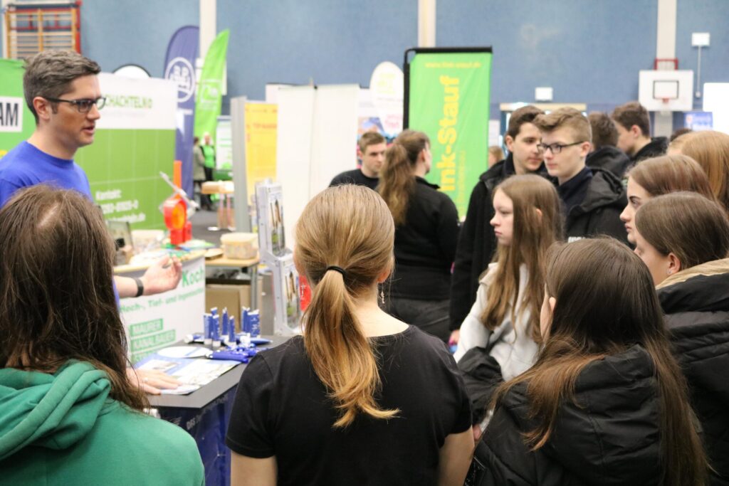 Ein Stand auf einer Messe mit einem Vortragenden und interessierten Jugendlichen.
