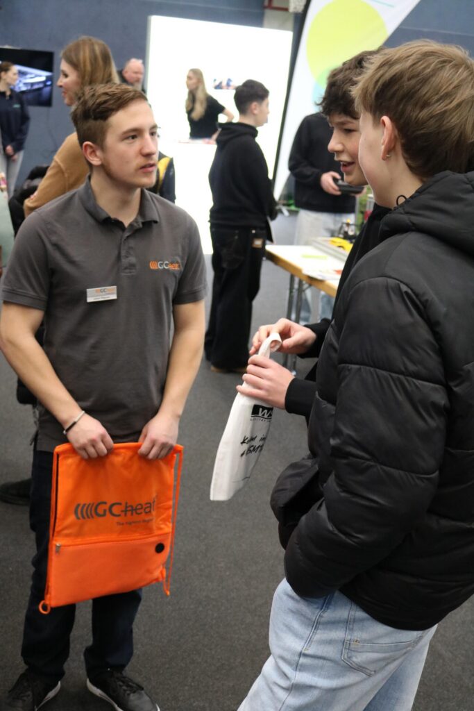 Junge Leute sprechen an einem Informationsstand, einer hält eine orange Tasche.
