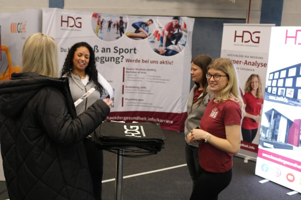 Messestand mit mehreren Personen, die Informationen austauschen und Broschüren halten.