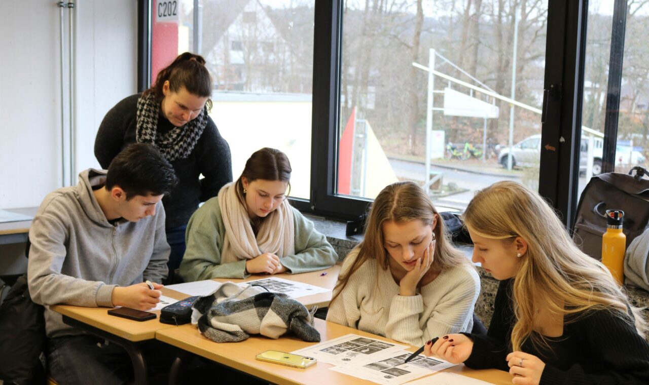 Schüler arbeiten an einem Tisch, während eine Lehrerin ihnen hilft.