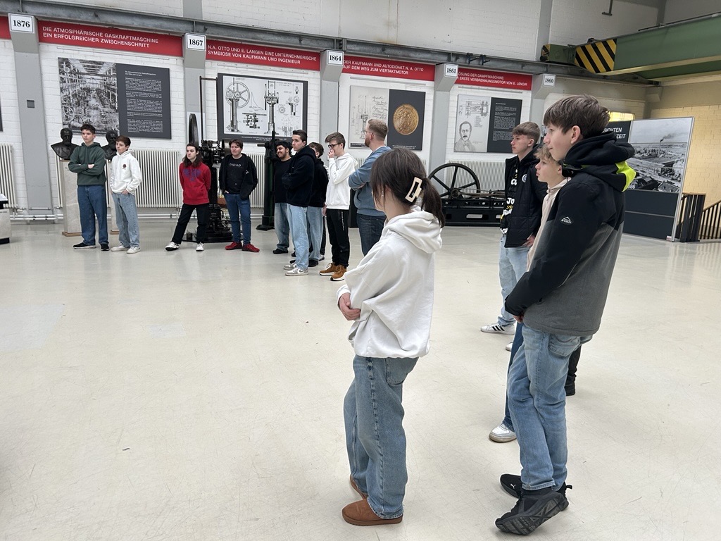 Schülerinnen und Schüler stehen in einem Raum und hören einer Präsentation zu.