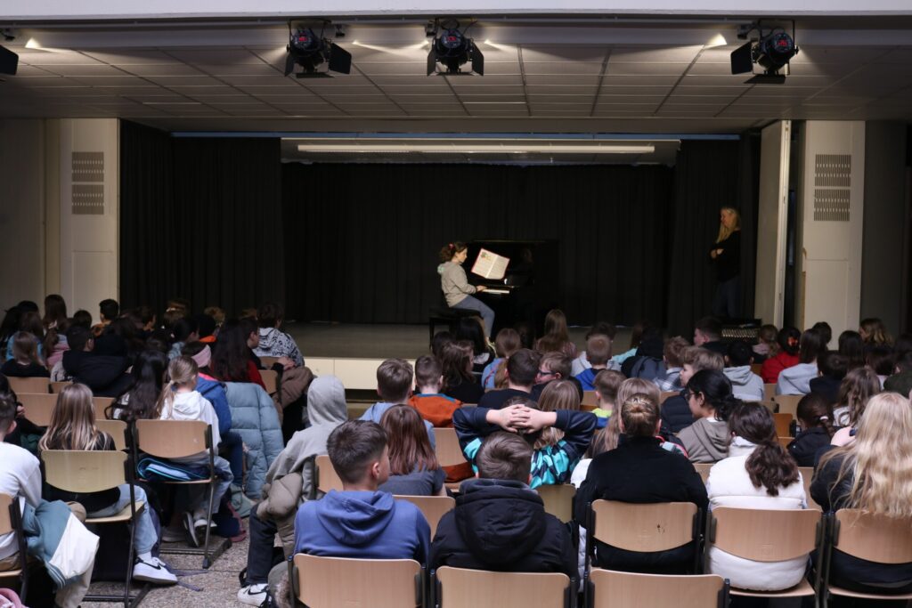 Schüler spielen Klavier auf einer Bühne vor einem Publikum in einem Raum.