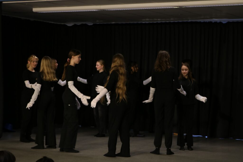 Schülerinnen und Schüler in schwarzen Kostümen mit weißen Handschuhen auf der Bühne.