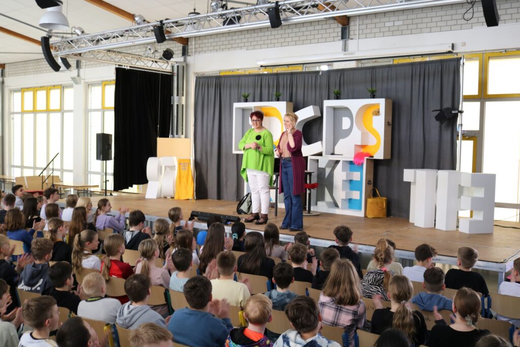 Schüler sitzen auf Stühlen, während zwei Personen auf einer Bühne sprechen.
