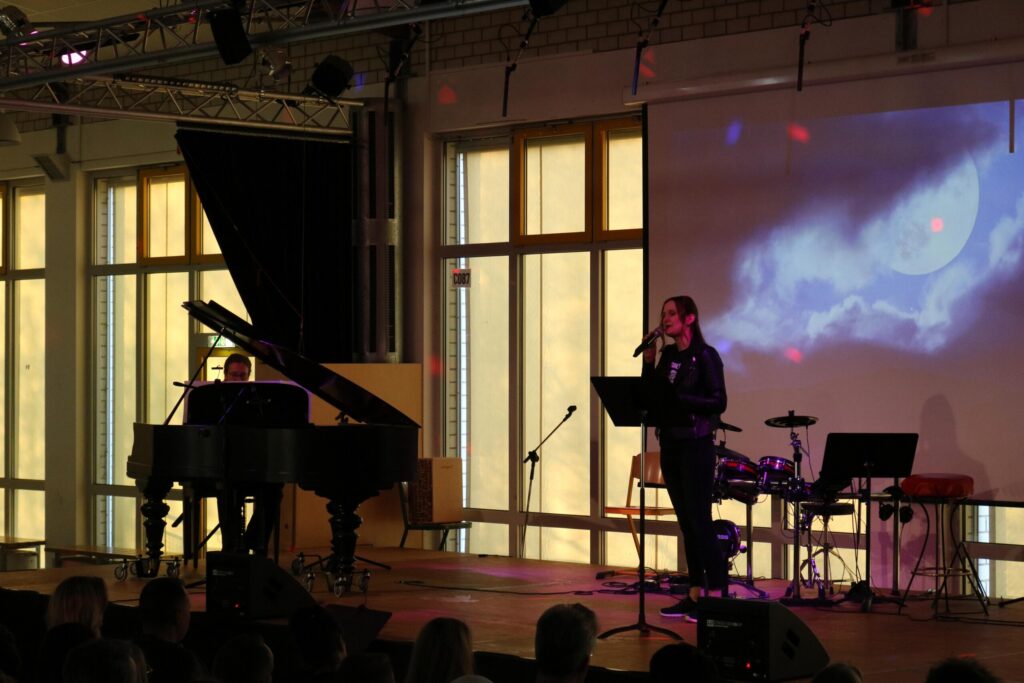 Schülerin singt am Mikrofon, Pianist spielt Klavier, Bühnenbeleuchtung vorhanden.