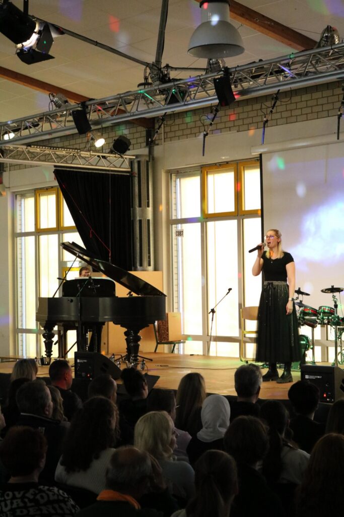 Schülerin singt auf der Bühne, begleitet von einem Pianisten. Publikum schaut zu.