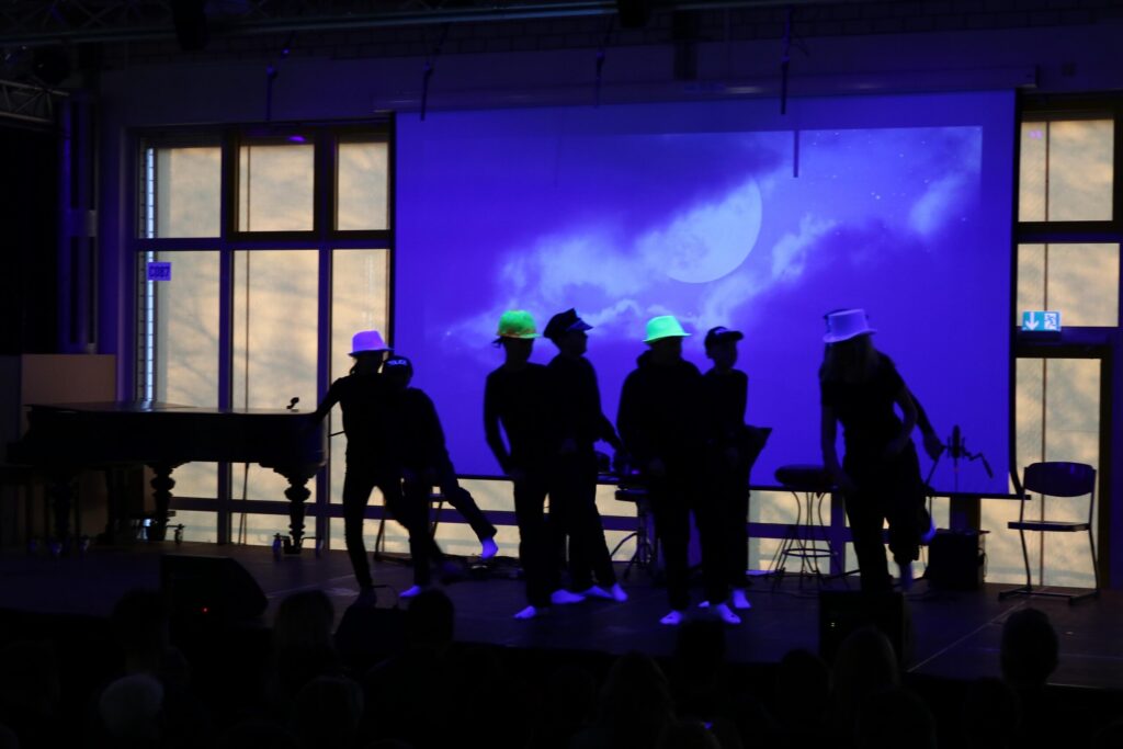 Schatten von Künstlern auf der Bühne mit Neonhüten vor einem projizierten Hintergrund.