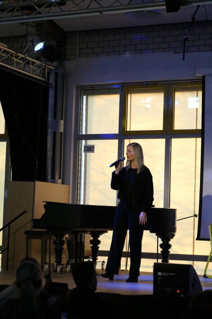Frau mit Mikrofon steht auf der Bühne vor einem Klavier.