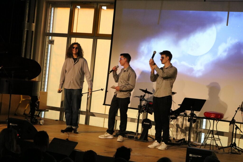 Drei Schüler singen auf einer Bühne vor einem Publikum, Instrumente im Hintergrund.