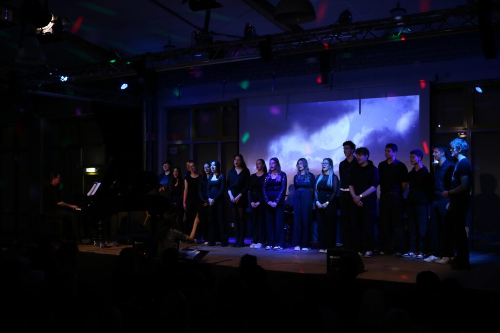 Schüler singen auf einer Bühne, im Hintergrund ein Klavier und Lichtspiele.