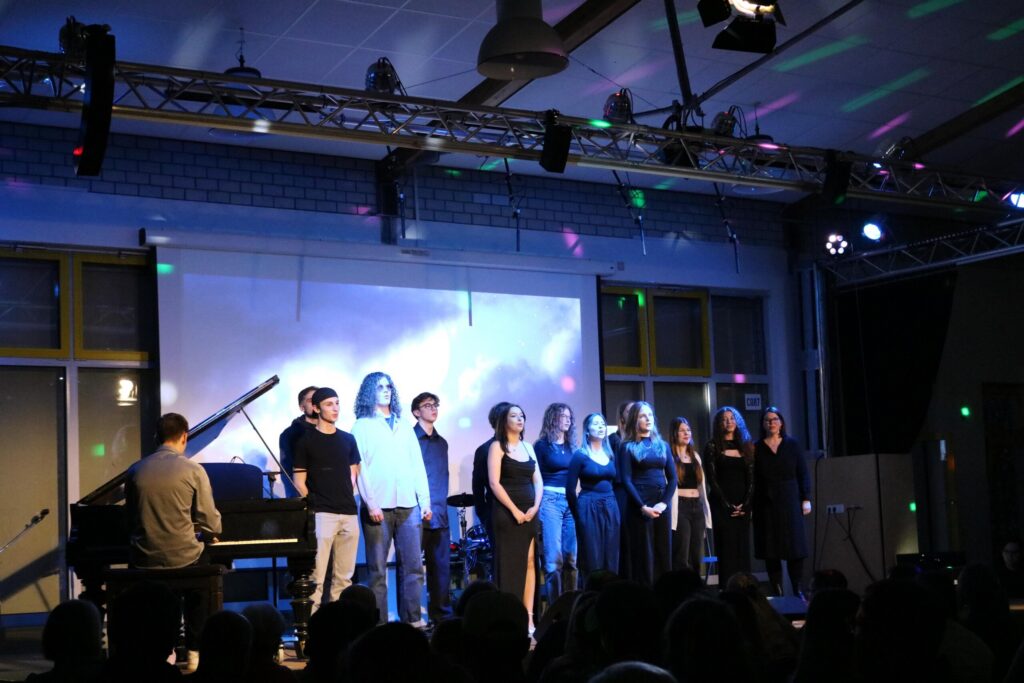 Auf der Bühne stehen Sängerinnen und Sänger, im Hintergrund ein Klavier. Lichtspiel.
