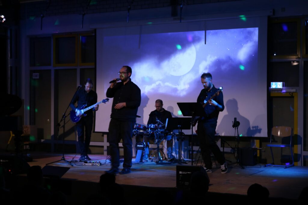 Band spielt auf der Bühne vor einer Projektion mit Wolken und farbigen Lichtern.