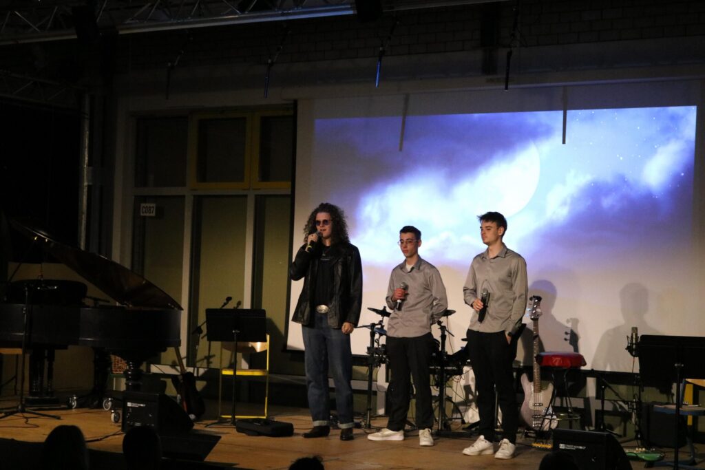 Drei Personen auf der Bühne, Hintergrund mit Projektion und Musikinstrumenten.