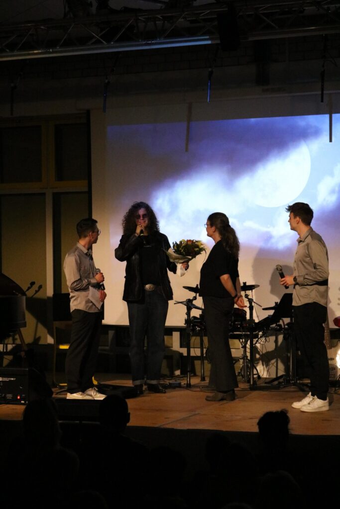 Eine Person hält Blumen auf einer Bühne, umgeben von zwei anderen Personen und Musikern.