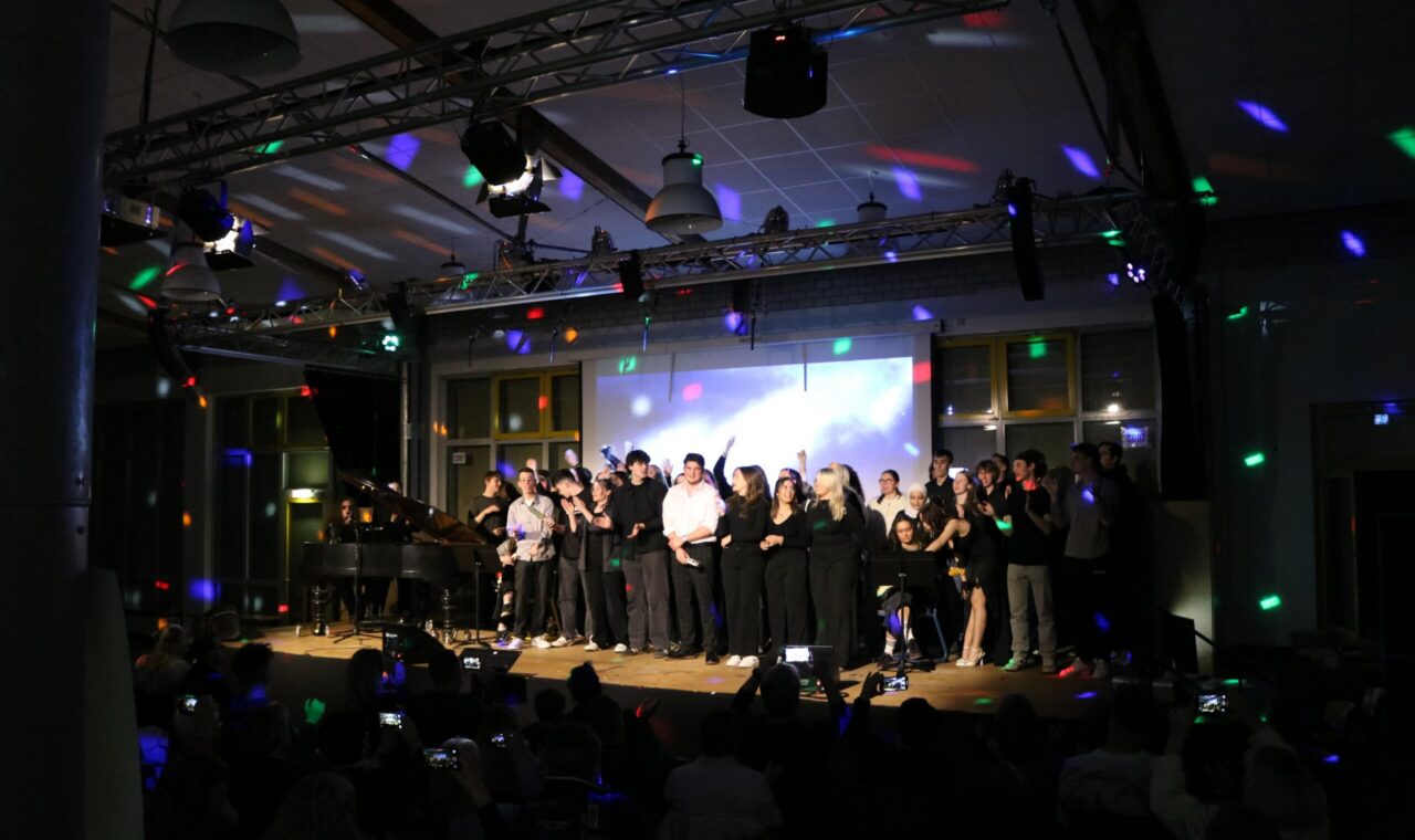 Schüler singen auf der Bühne, buntes Lichtspiel im Hintergrund. Publikum applaudiert.