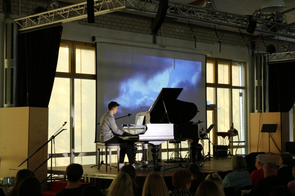 Ein Pianist spielt auf einem weißen Klavier vor einer großen Leinwand.