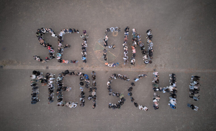 Menschen bilden den Schriftzug „SEI EIN MENSCH“ aus der Vogelperspektive.