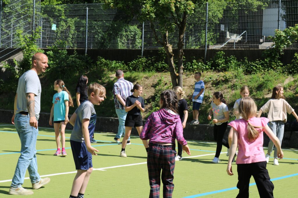 Kinder spielen auf einem Sportplatz, Aufsichtsperson beobachtet das Geschehen.