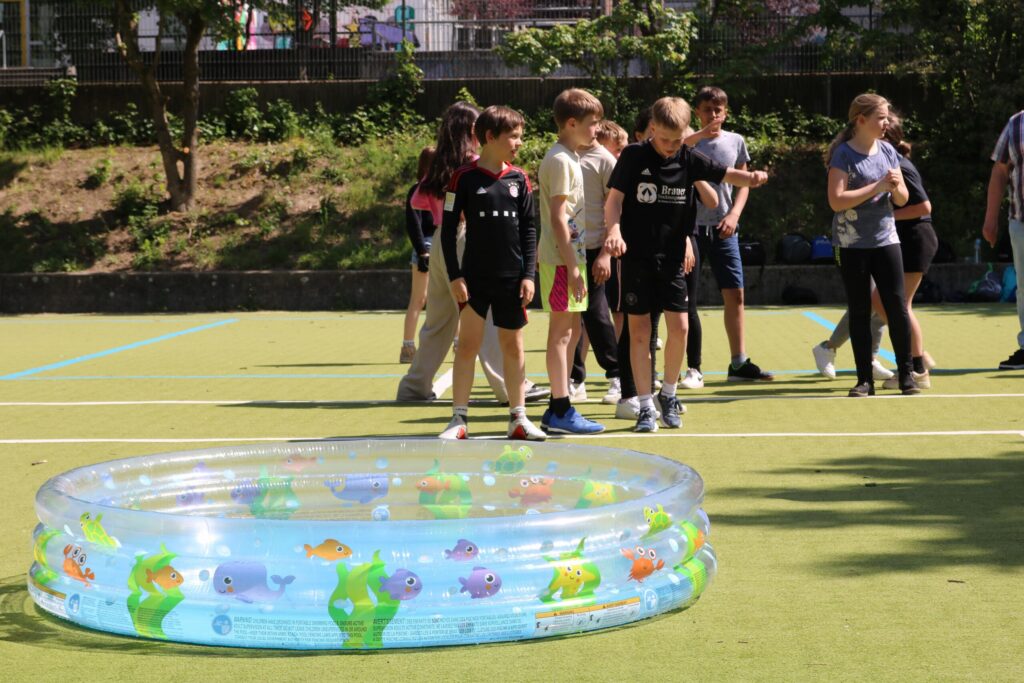 Kinder stehen um ein aufblasbares Planschbecken auf einem Sportplatz.