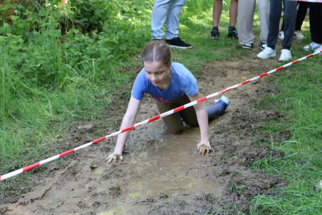 Mädchen kriecht durch eine matschige Stelle im Freien, Zuschauer stehen daneben.