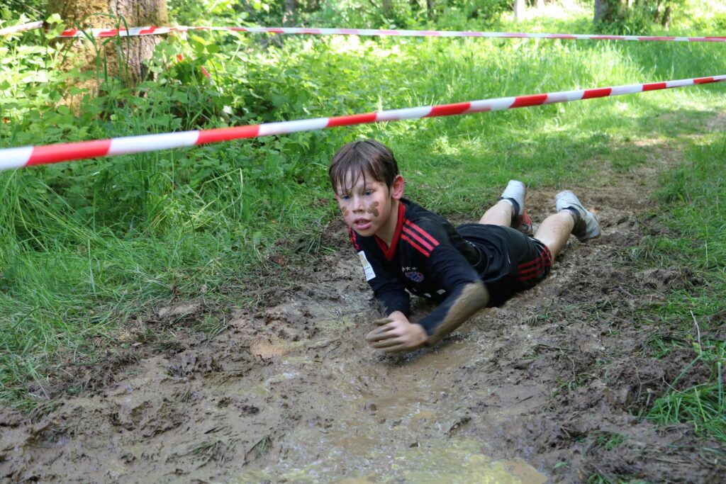 Ein Junge liegt im Schlamm und kriecht unter einem Absperrband hindurch.