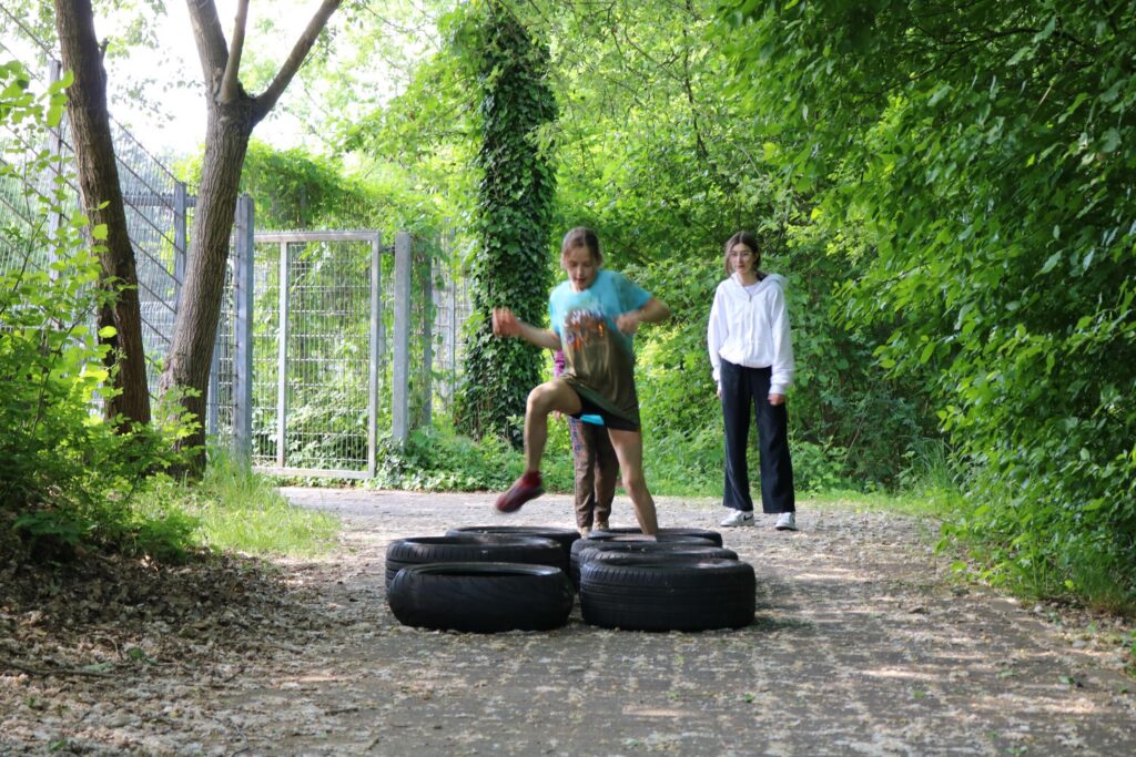 Kinder springen über Autoreifen auf einem Waldweg. Eine Erwachsene beobachtet.