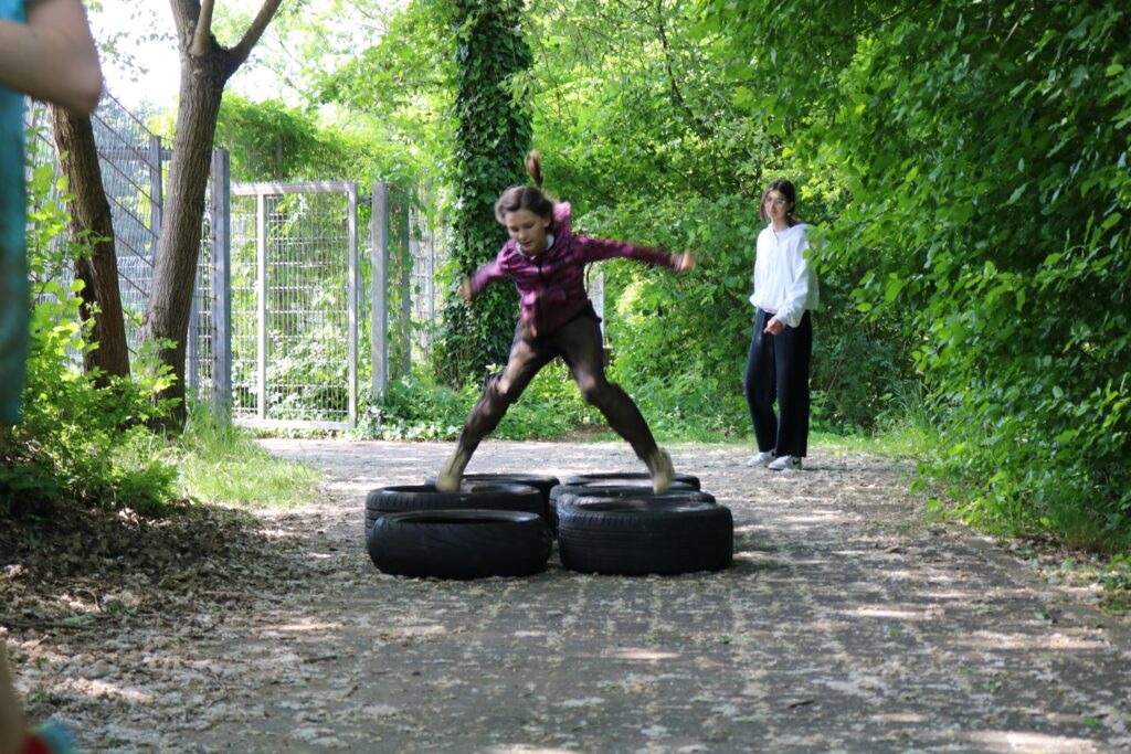 Ein Kind springt über Reifen auf einem Waldweg, während eine weitere Person zuschaut.