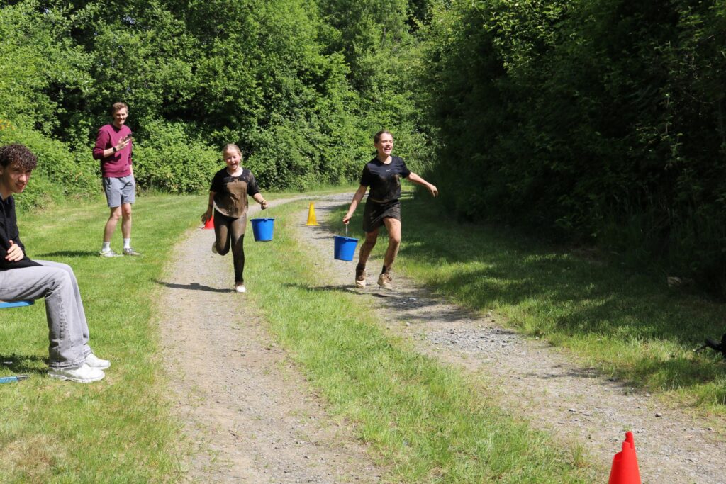 Zwei Kinder rennen mit Eimern auf einem Feldweg, während ein Erwachsener zuschaut.