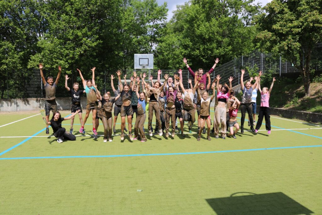 Gruppenselfie von vielen Personen auf einem Sportplatz, alle jubeln und haben Spaß.