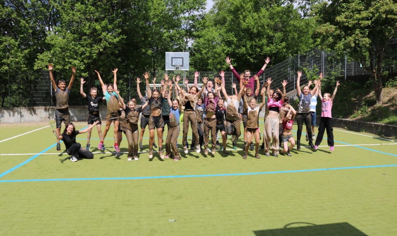 Gruppenselfie von vielen Personen auf einem Sportplatz, alle jubeln und haben Spaß.