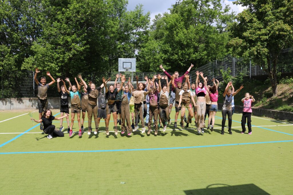 Gruppe von Jugendlichen in sportlicher Kleidung posiert auf einem Platz, alle zeigen Freude.