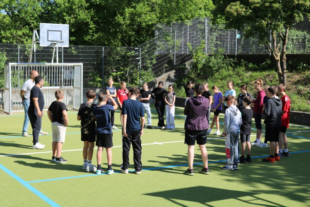 Gruppe von Schülern versammelt auf dem Sportplatz, Lehrer erklärt eine Übung.
