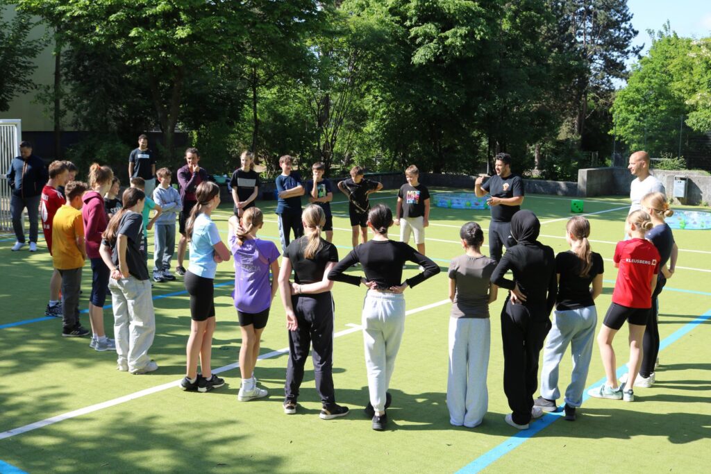 Schüler stehen in einem Kreis auf einem Sportplatz und hören einem Trainer zu.