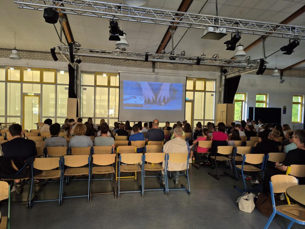 Blick auf Publikum in einem Raum vor einer Leinwand mit einer Präsentation.