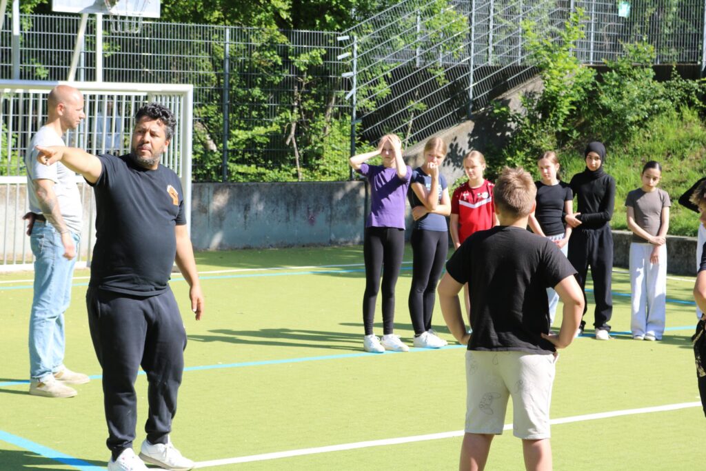 Eine Gruppe von Jugendlichen steht auf einem Sportplatz, ein Trainer erklärt etwas.