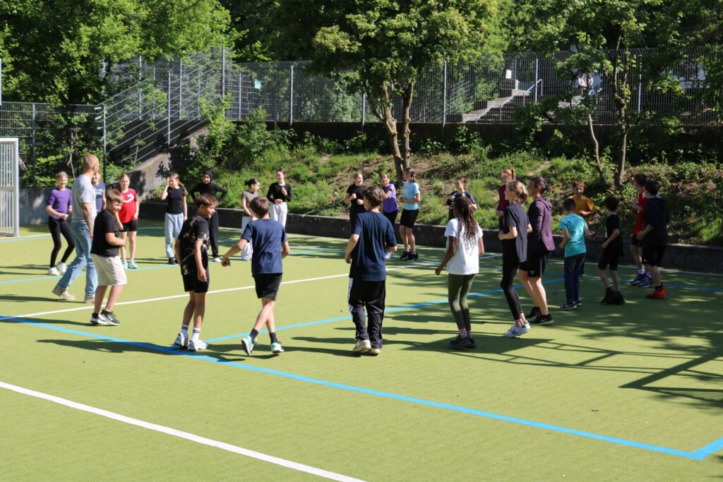 Schülerinnen und Schüler spielen auf einem Sportplatz bei sonnigem Wetter.