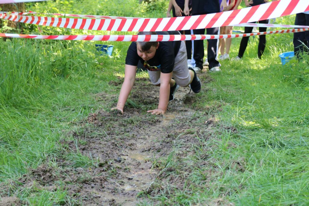 Schüler kriecht unter Absperrband durch einen matschigen Bereich im Freien.