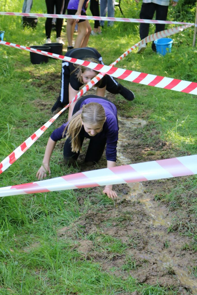 Schülerinnen kriechen unter Absperrbändern hindurch auf einem matschigen Boden.