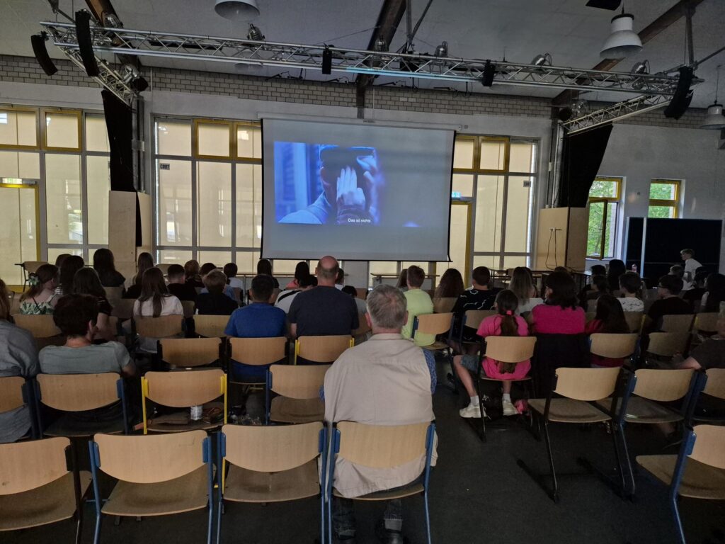 Schüler und Erwachsene sitzen im Raum vor einer Leinwand.