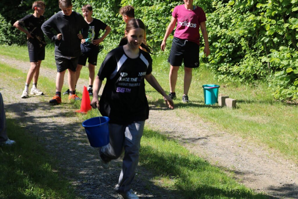 Schülerin trägt Eimer und läuft auf einem Weg, im Hintergrund stehen andere Schüler.