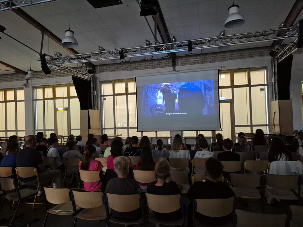 Eine Gruppe von Menschen sitzt in einem Raum und schaut einen Film auf einer Leinwand.