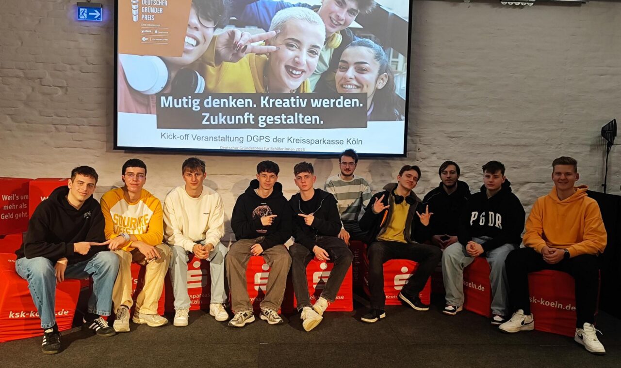 Gruppe junger Menschen sitzt auf einer Bühne mit einem Präsentationsbild im Hintergrund.