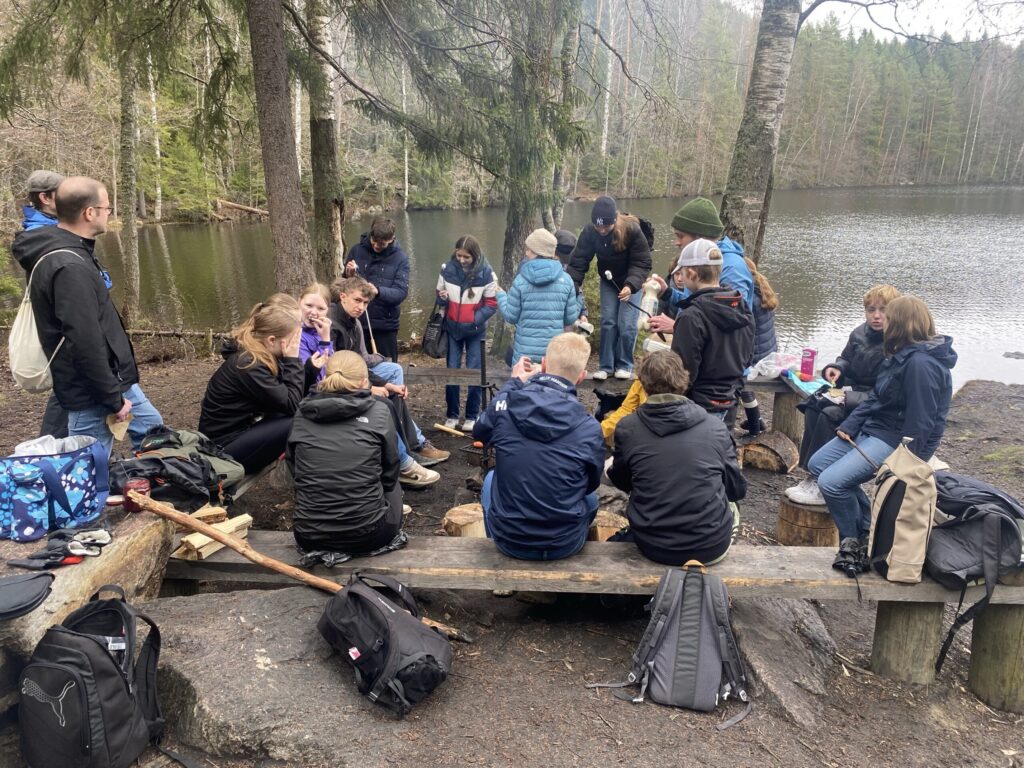 Gruppe von Schülerinnen und Schülern sitzt am Ufer eines Sees im Wald.