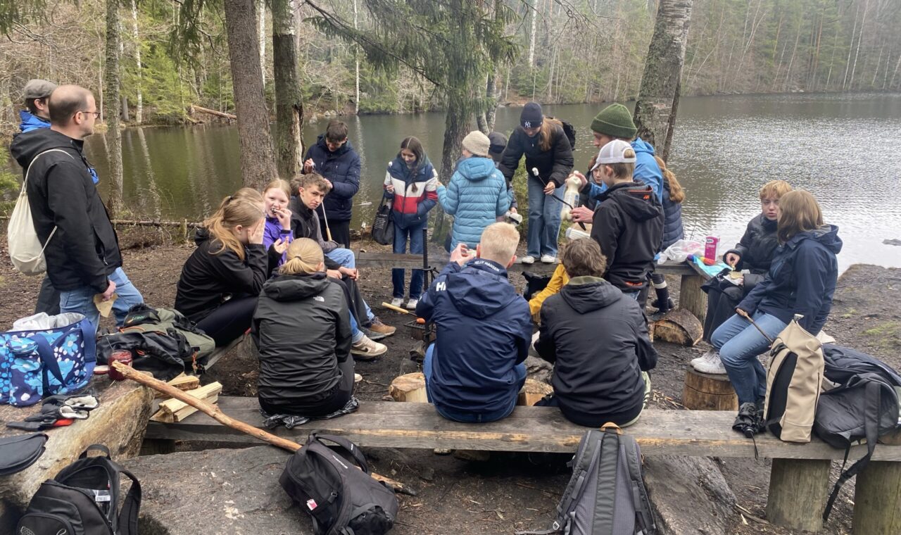 Gruppe von Schülerinnen und Schülern sitzt am Ufer eines Sees im Wald.