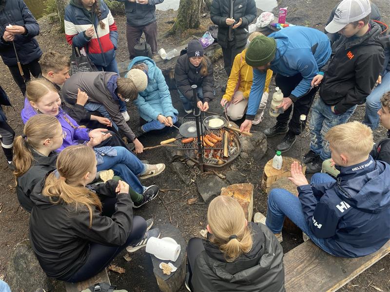 Gruppe von Schülern am Lagerfeuer, die Holzstäbe mit Essen halten.